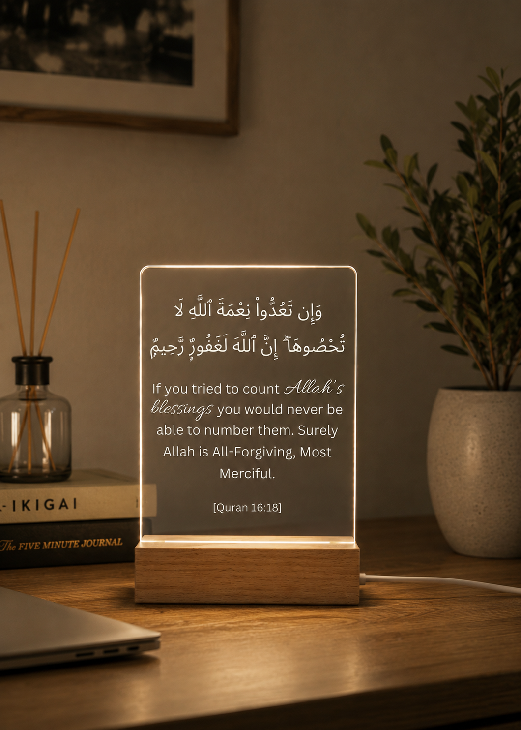 Decorative lamp with Arabic text on a wooden base, placed on a table with a plant and books in the background.