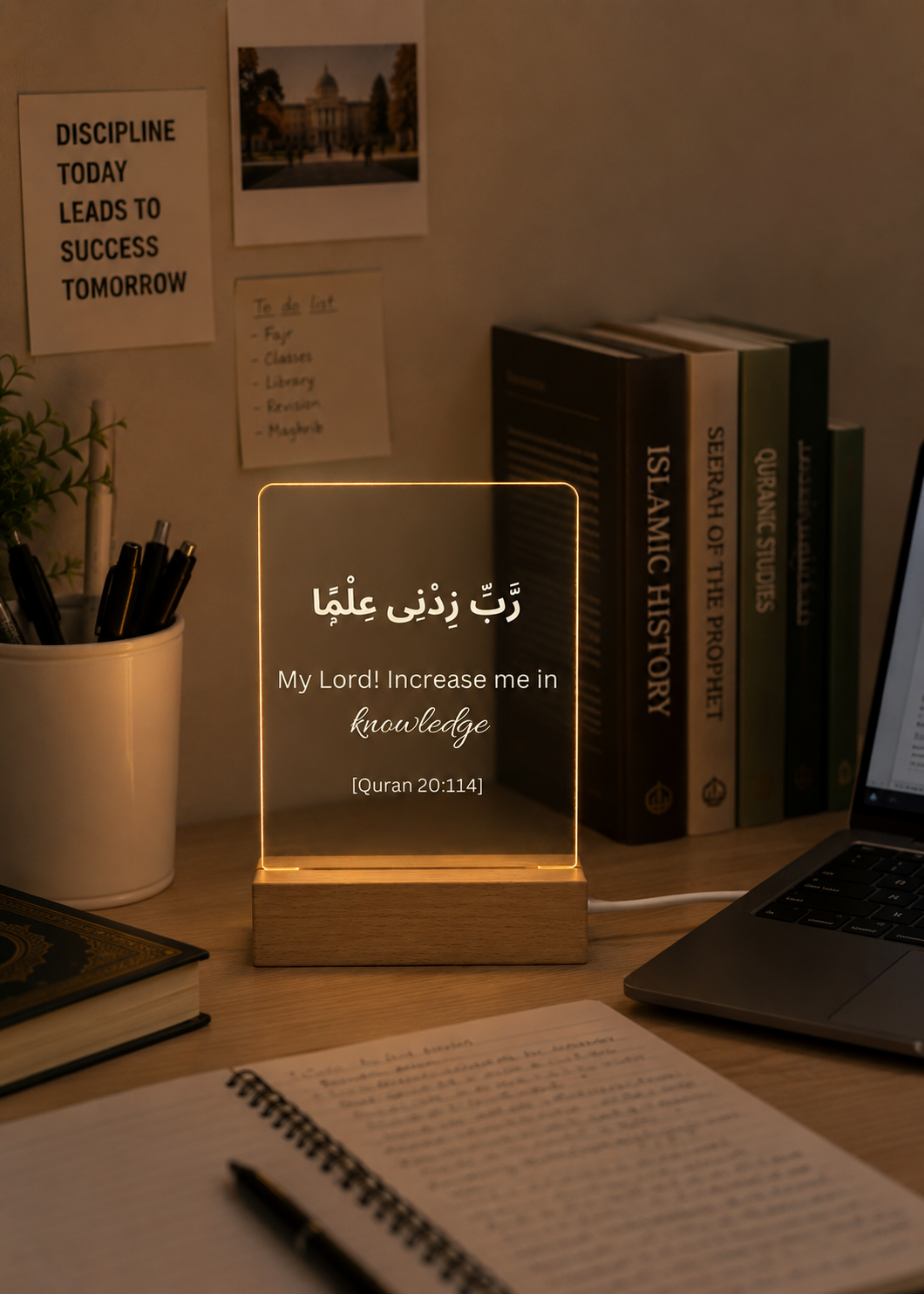 Desk setup with a glowing transparent lamp displaying Arabic text, books, and a laptop.
